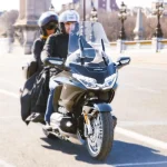 Un couple en moto Honda Gold Wing roule sur une route de ville, avec un paysage urbain en arrière-plan.