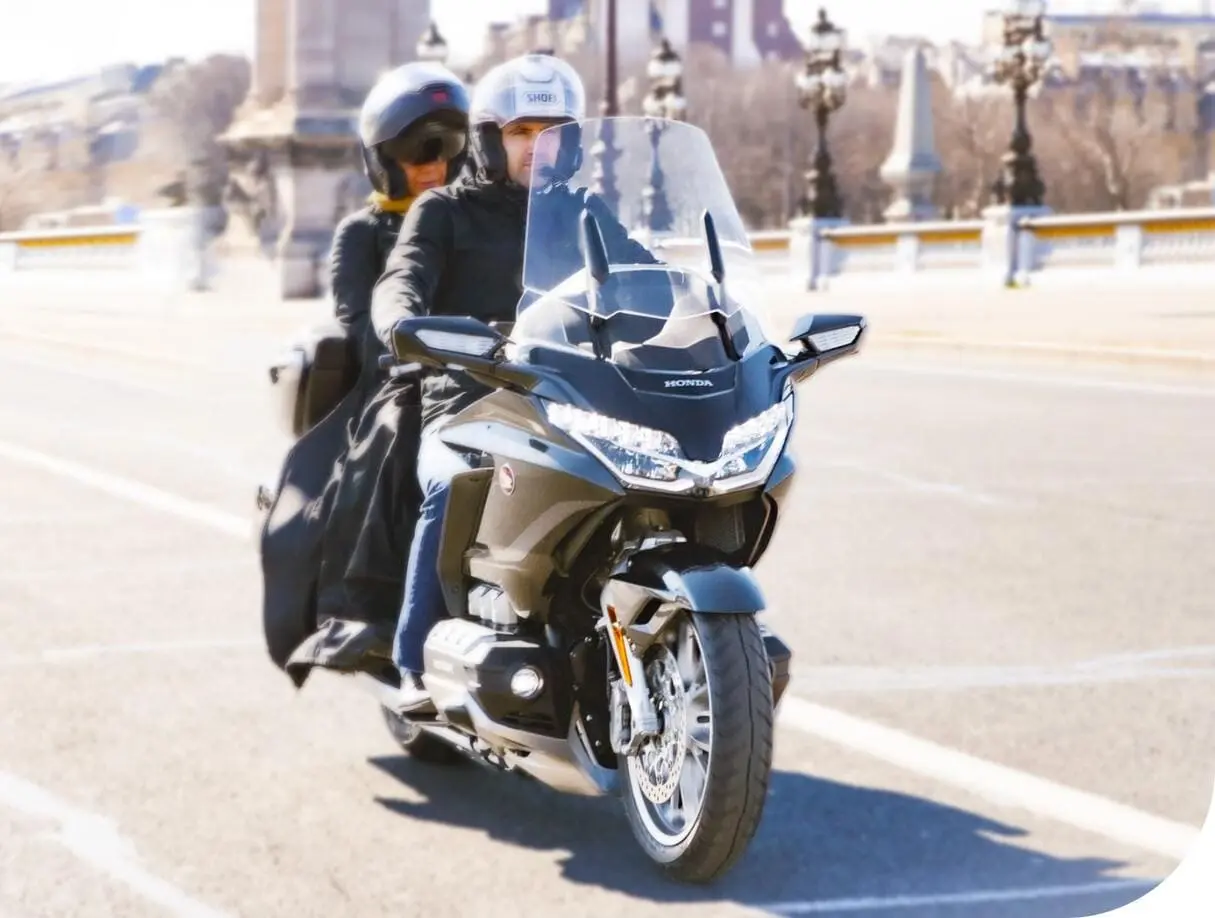 Un couple en moto Honda Gold Wing roule sur une route de ville, avec un paysage urbain en arrière-plan.