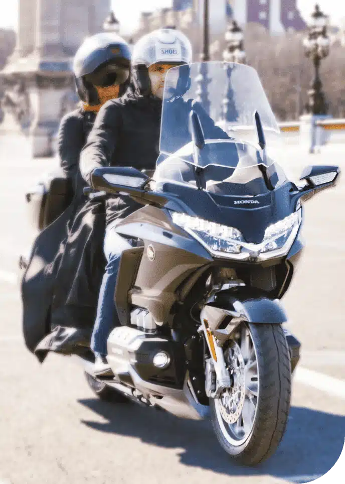 Un couple en moto Honda Gold Wing roule sur une route de ville, avec un paysage urbain en arrière-plan.