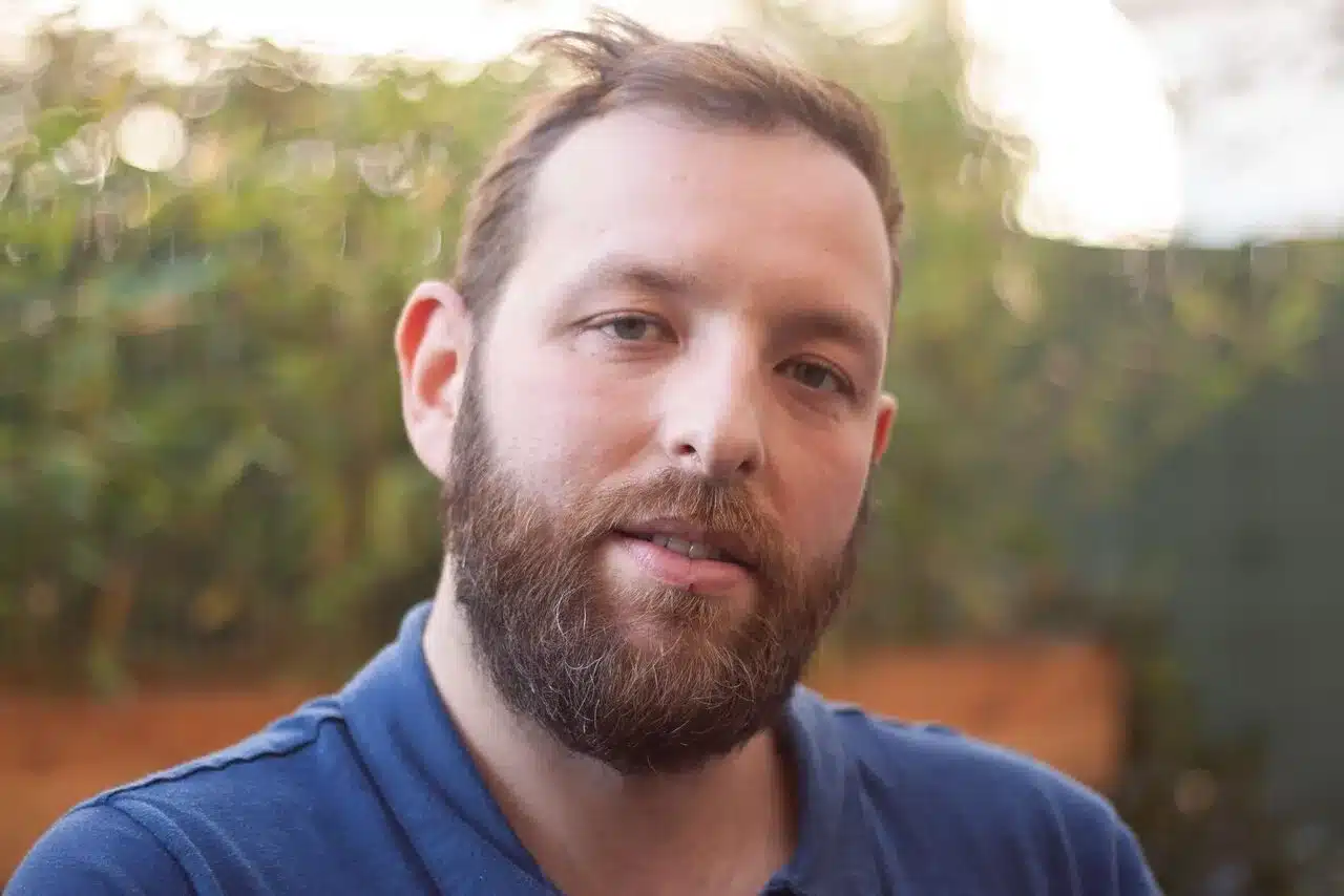 Un homme souriant et barbue. En arrière plan de la végétation.