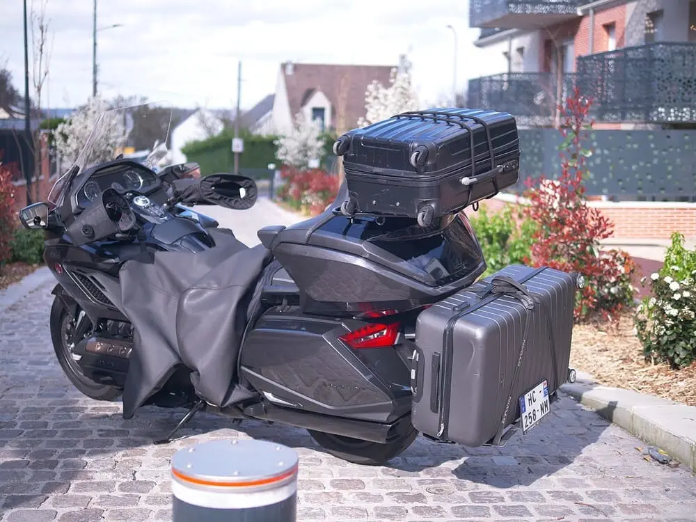 Une moto au premier plan, avec plusieurs valises. En arrière-plan des pavillons